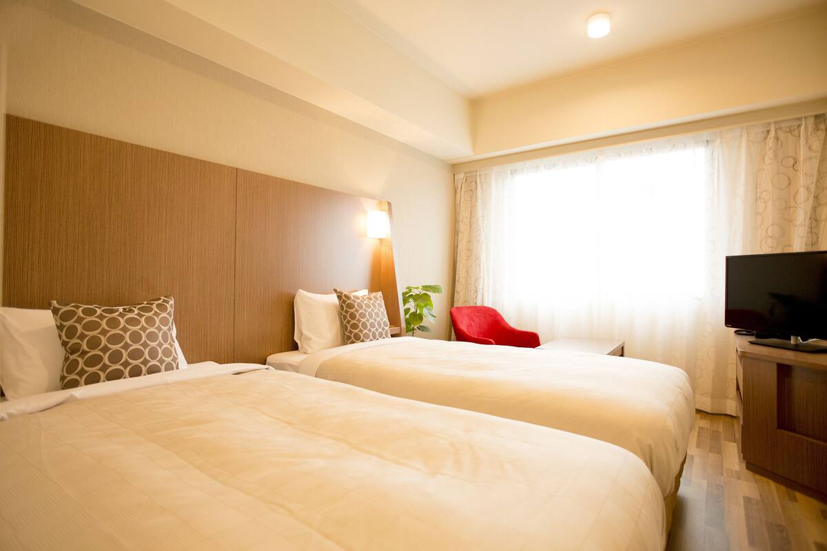 A hotel room with two beds, a TV, a red chair, and a window with curtains.