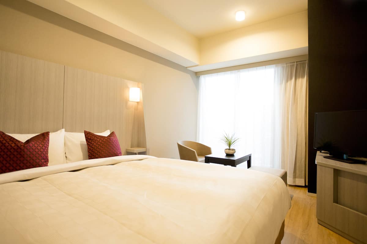 A hotel room with a large bed, a TV, a desk with a plant, and a window with curtains.