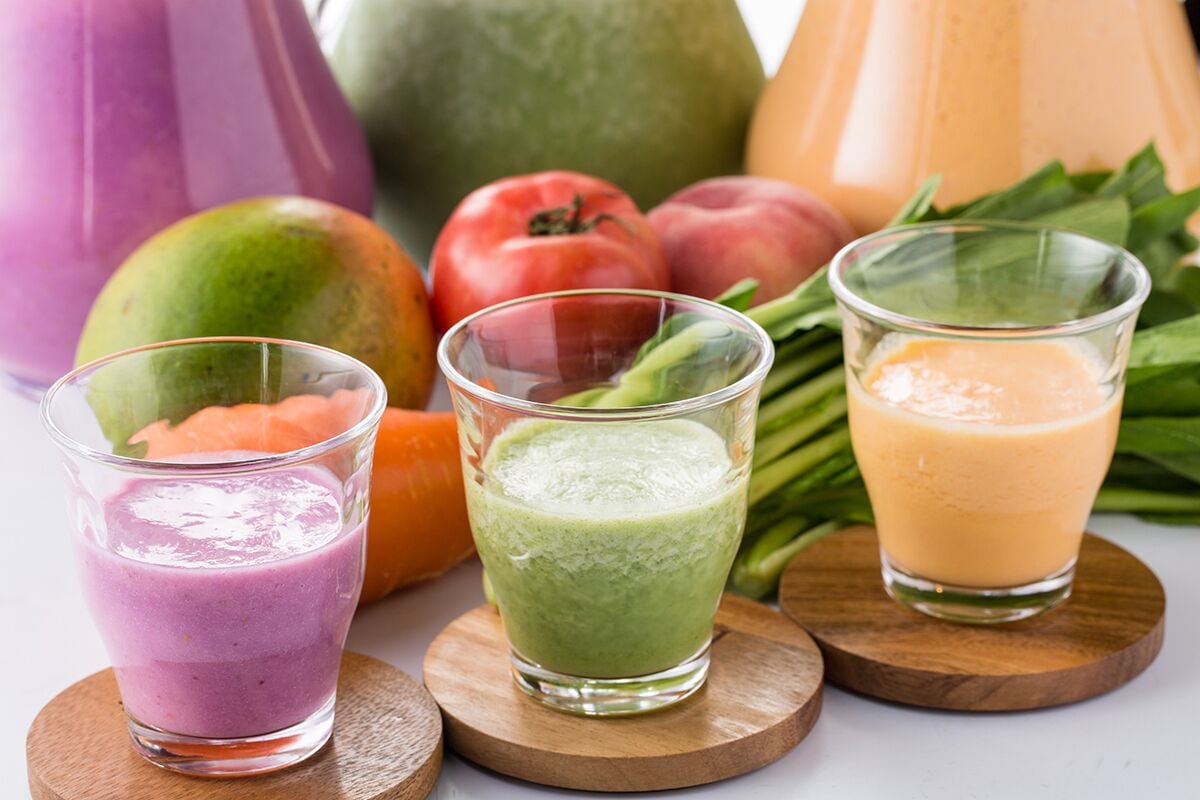 Three glasses of smoothie on wooden coasters with fresh fruits in the background.