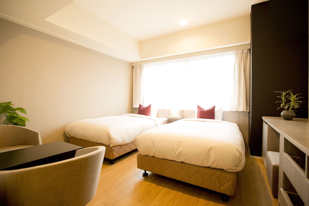 A hotel room with two beds, a desk, a chair, a potted plant, and a large window with curtains.