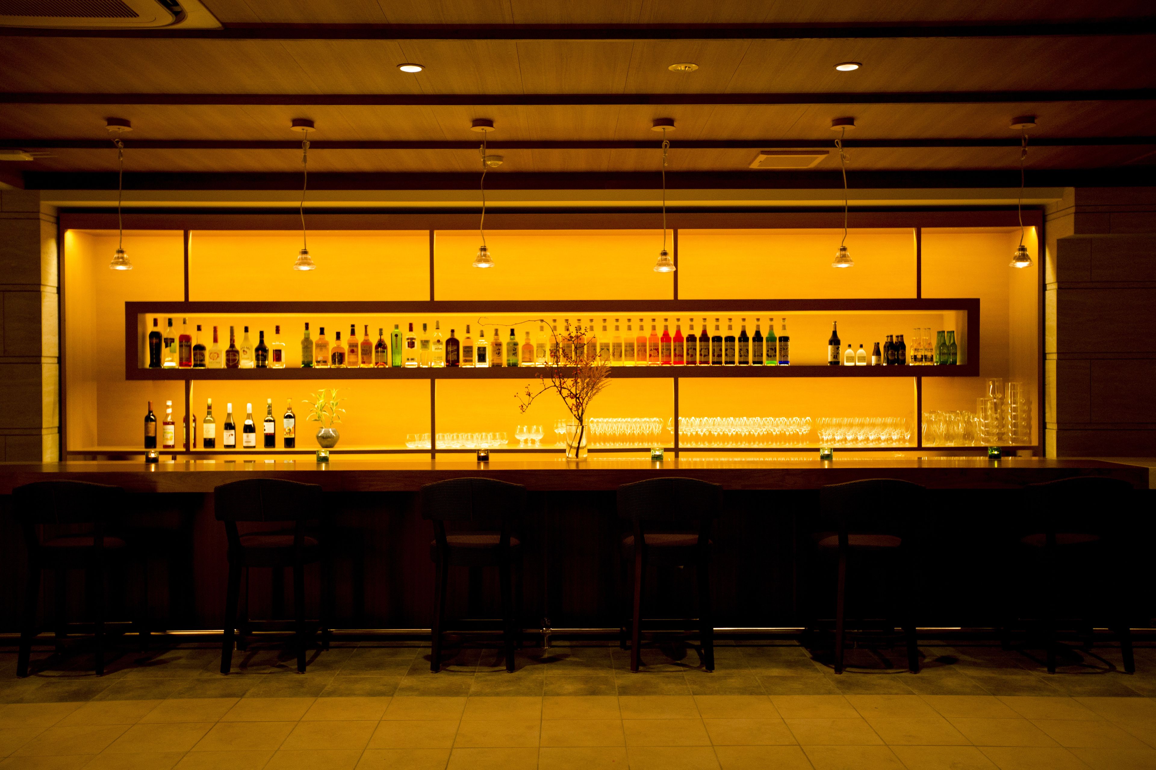 A bar area with multiple shelves of bottles and glasses, wooden bar stools, and hanging lights.