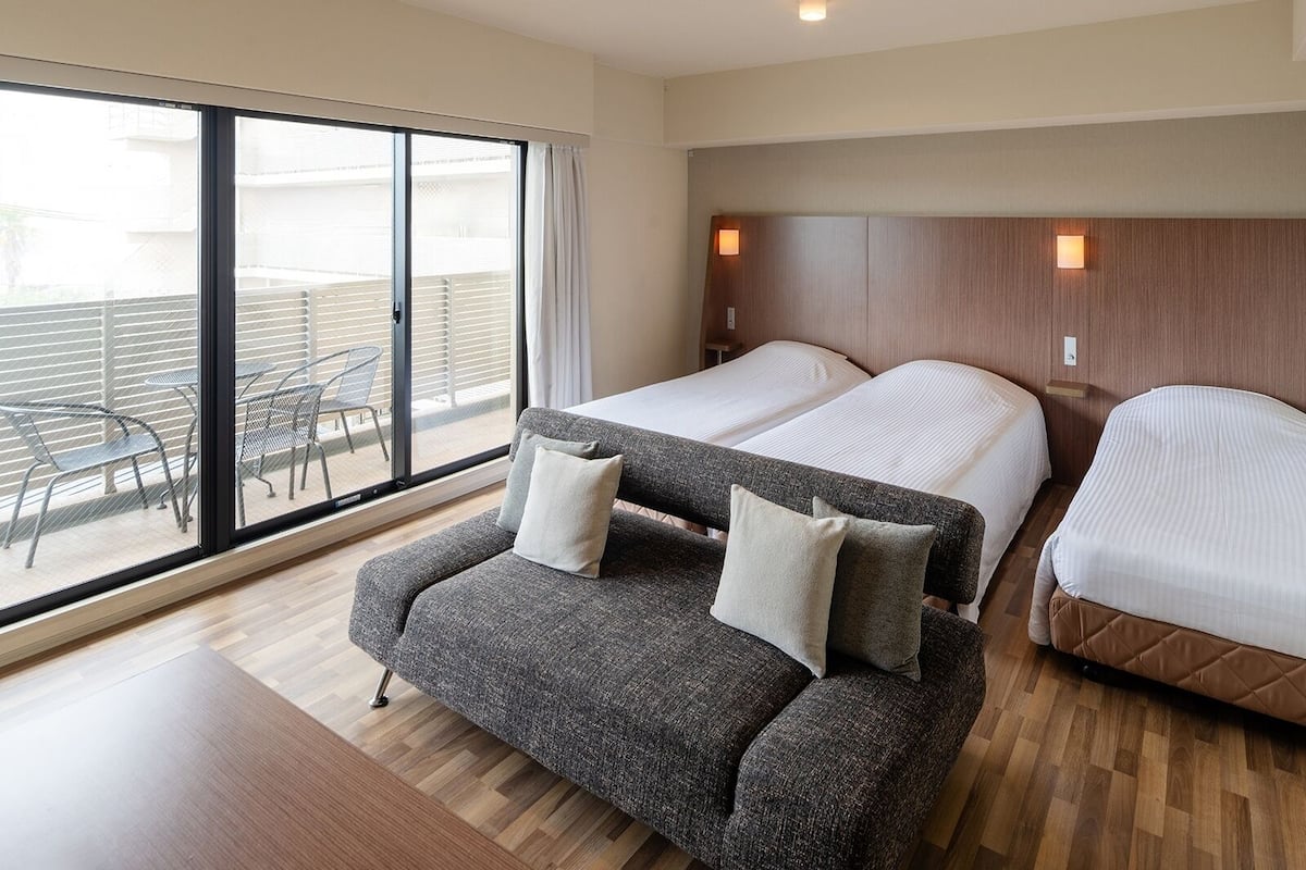 A hotel room with two beds, a sofa, and a balcony with outdoor furniture.