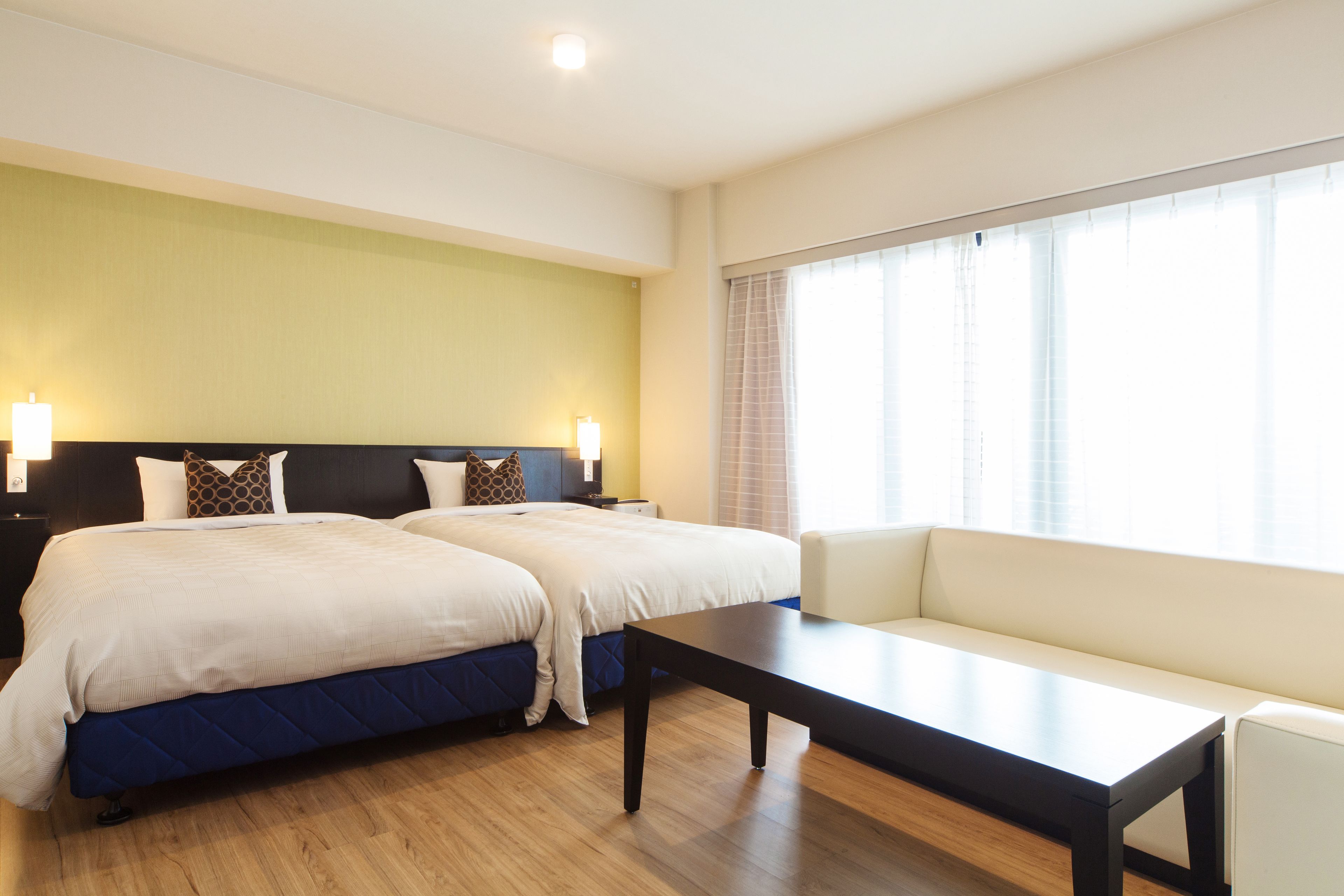 A hotel room with two beds, a sofa, a table, and a large window with curtains.