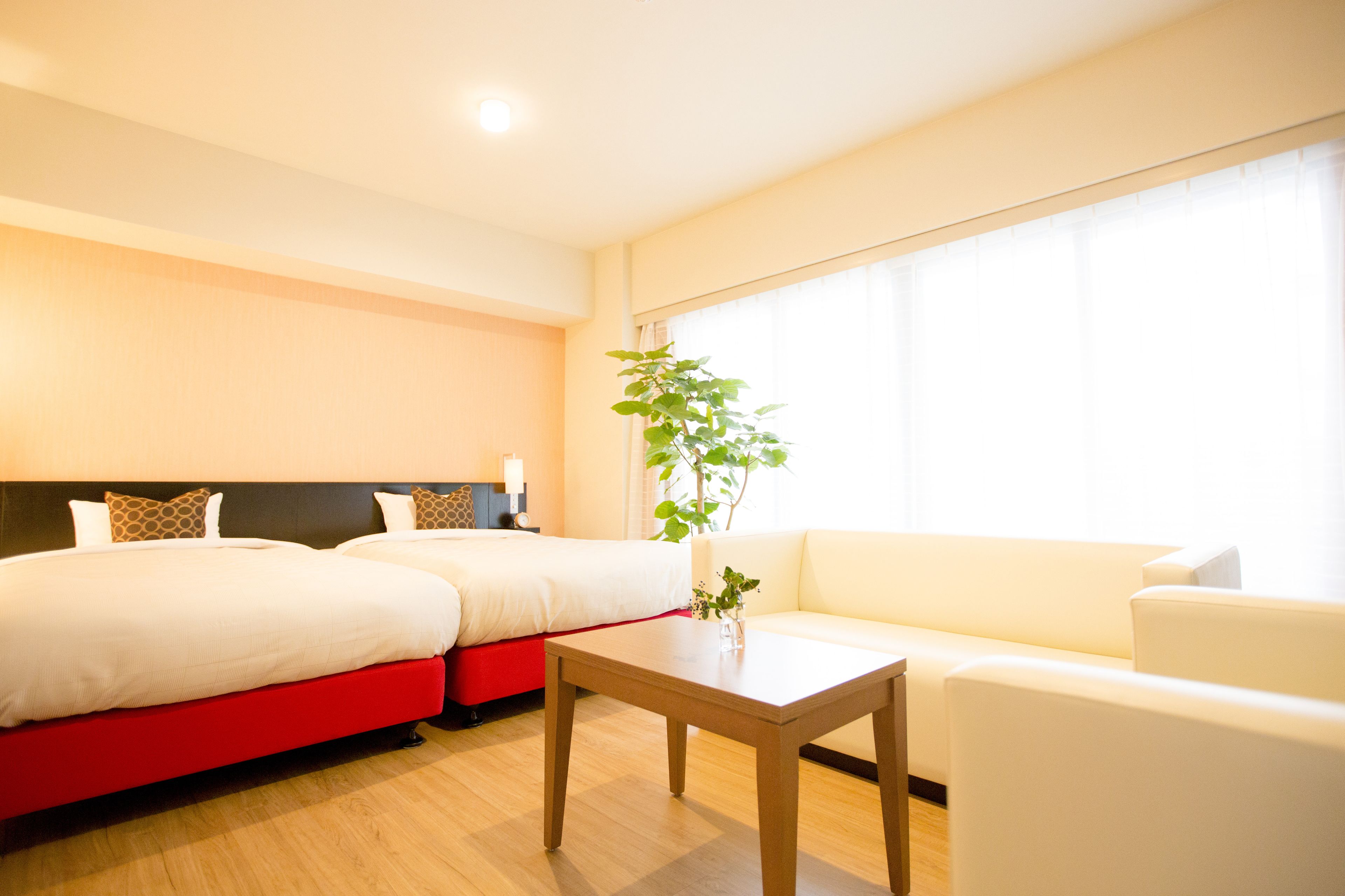 A hotel room with two beds, a red accent bed, a white sofa, a wooden table, and a large window with curtains.
