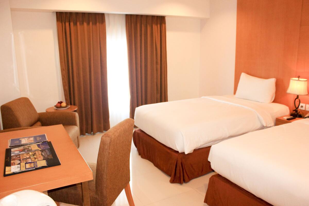 Habitación de hotel con dos camas, una mesita con una revista, una silla, una mesita de noche con una lámpara y una ventana con cortinas.