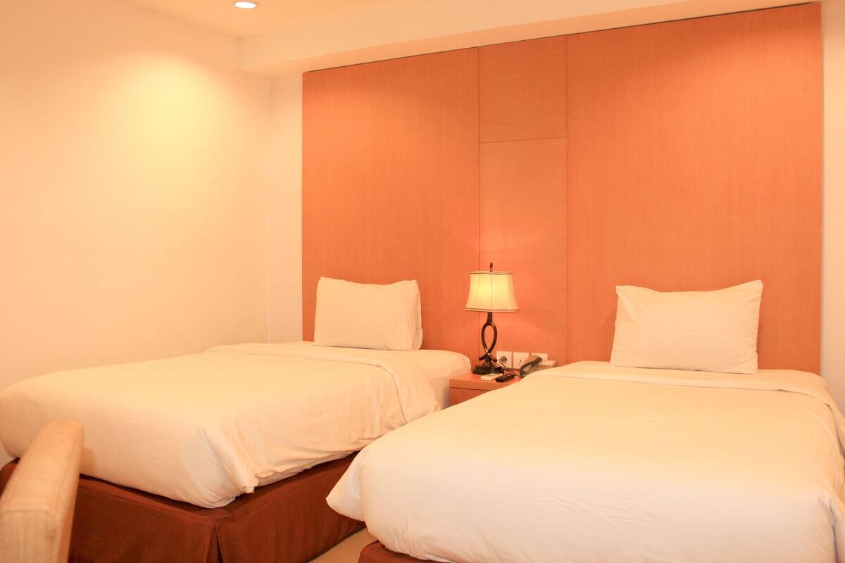 Habitación de hotel con dos camas, una mesita de noche con lámpara y cabecero de madera.