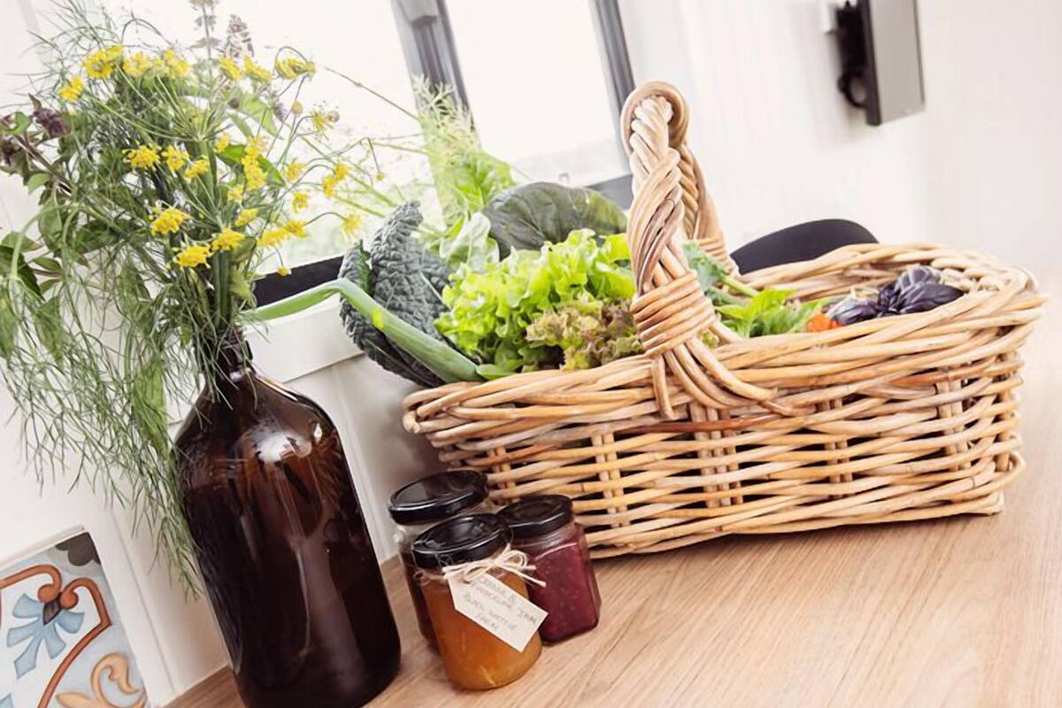 Un panier en osier rempli de légumes et d’herbes aromatiques, un vase en verre brun orné de fleurs jaunes et deux bocaux de conserves.