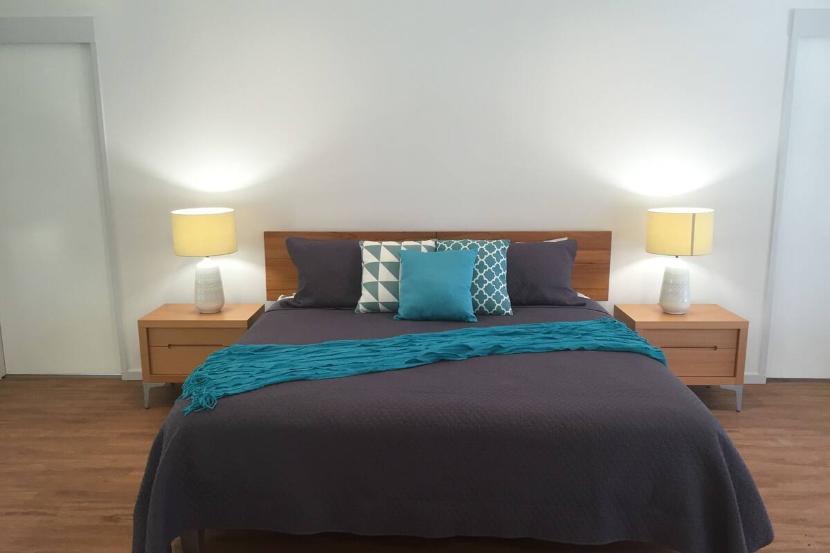 Un lit bien fait, recouvert d’une couvre-lit bleu foncé, agrémenté d’un plaid turquoise et de plusieurs coussins aux motifs variés. Deux tables de chevet, sur lesquelles reposent des lampes et des vases décoratifs, se trouvent de part et d’autre du lit. La pièce est dotée d’un plancher en bois et de murs blancs.