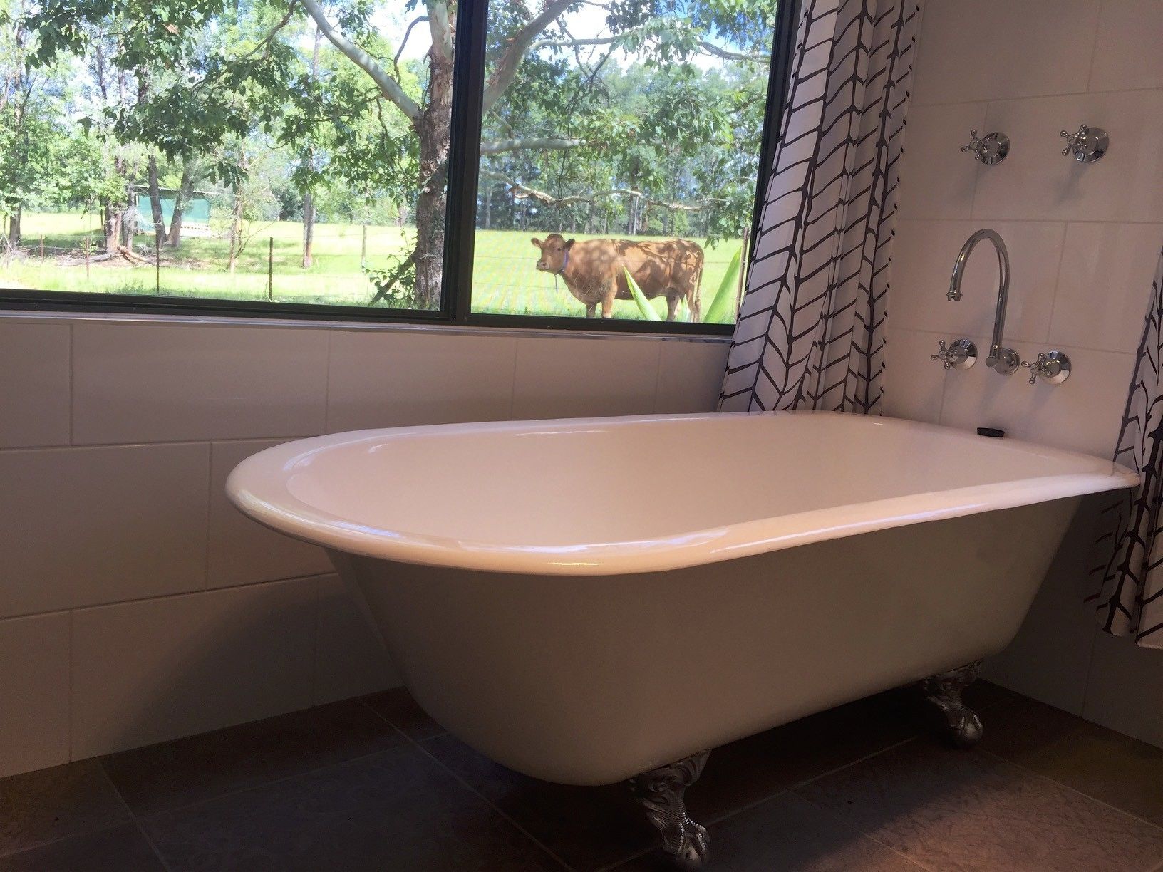 A bathroom with a freestanding bathtub, a window with a view of a cow in a field, and a shower area with a patterned curtain.