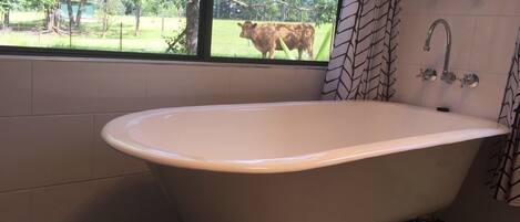 A bathroom with a freestanding bathtub, a window with a view of a cow in a field, and a shower area with a patterned curtain.