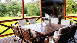 Een overdekt terras met een houten tafel en stoelen, een barbecue en uitzicht op een groen landschap.