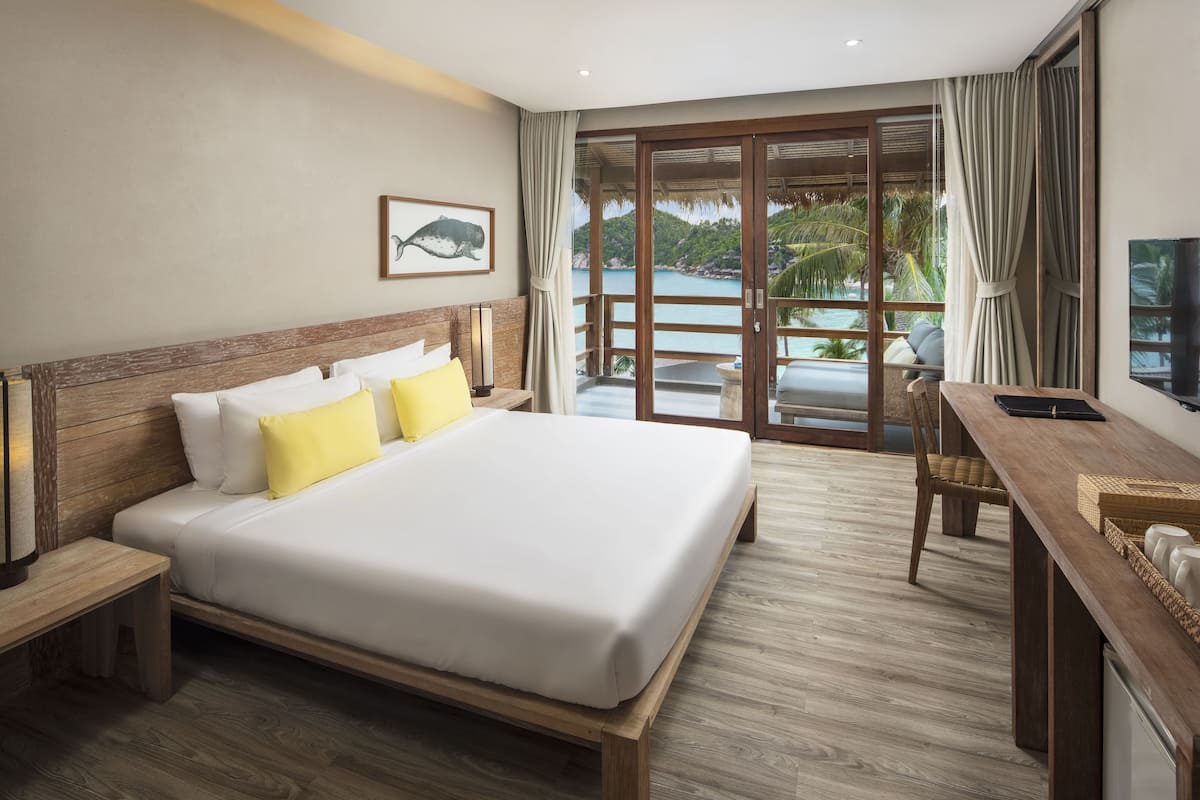 Une chambre d’hôtel avec un grand lit, une tête de lit en bois et un bureau avec une chaise. Une grande fenêtre offre une vue sur l’océan et les palmiers.