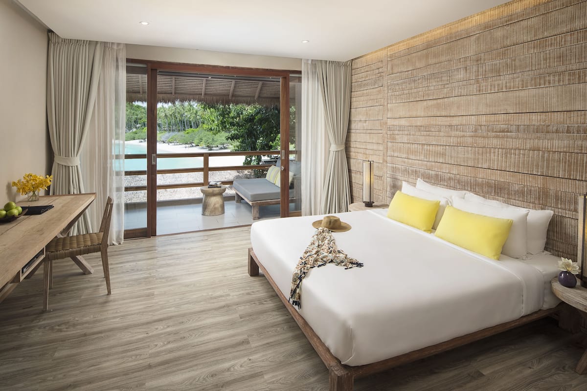Une chambre d’hôtel avec un grand lit, un plancher en bois, une table à manger avec des chaises, une vue sur la plage et un balcon avec une table et des chaises.