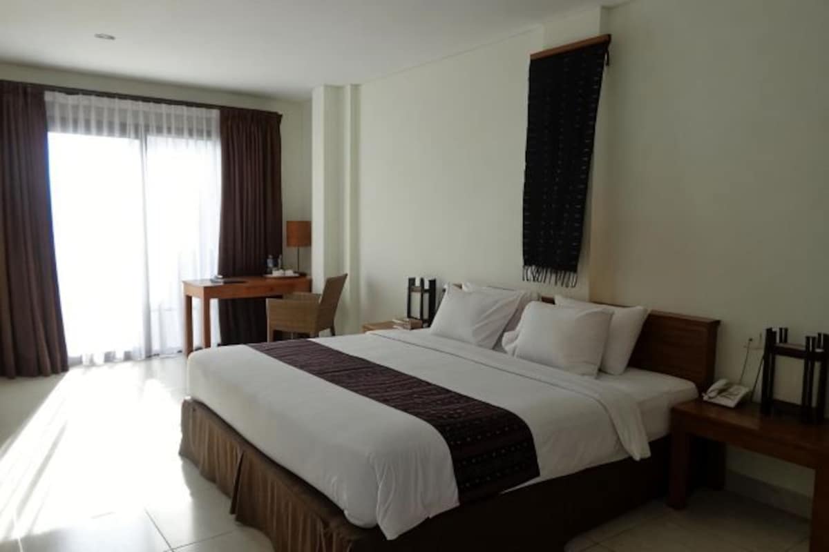 A hotel room with a large bed, a desk, a chair, a nightstand, and a window with curtains.