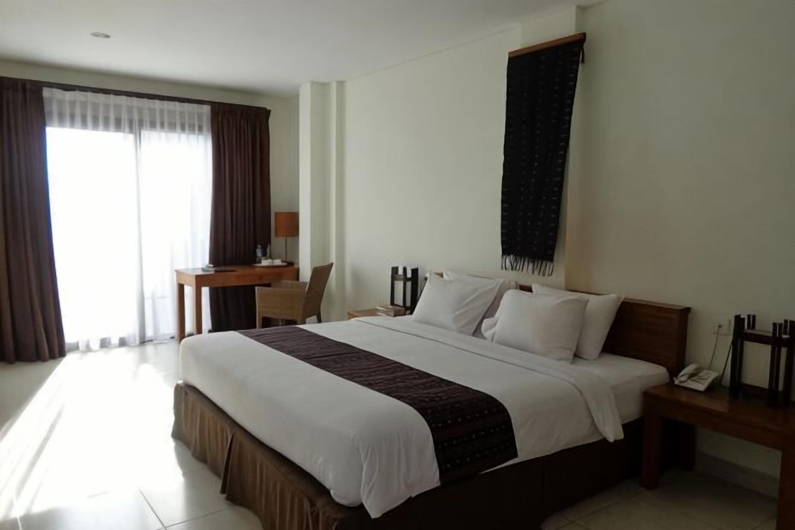 A hotel room with a large bed, a desk, a chair, a nightstand, and a window with curtains.