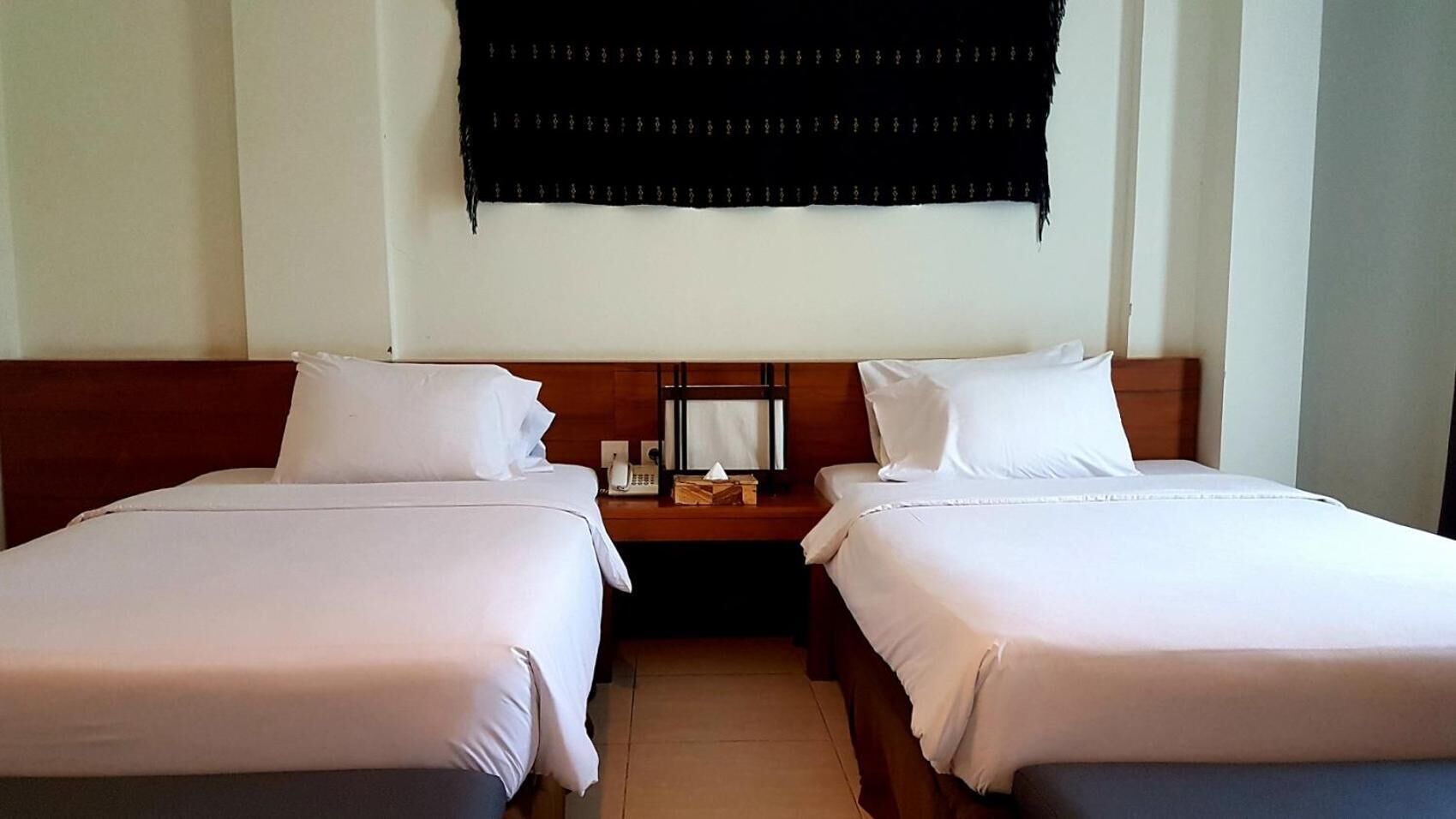 A hotel room with two single beds, a wooden headboard, a nightstand with a lamp, and a black curtain on the window.