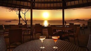 Un restaurant doté d’une terrasse offrant une vue imprenable sur le coucher du soleil. Les tables sont disposées avec des chaises, et un verre contenant une boisson est posé sur l’une d’elles.