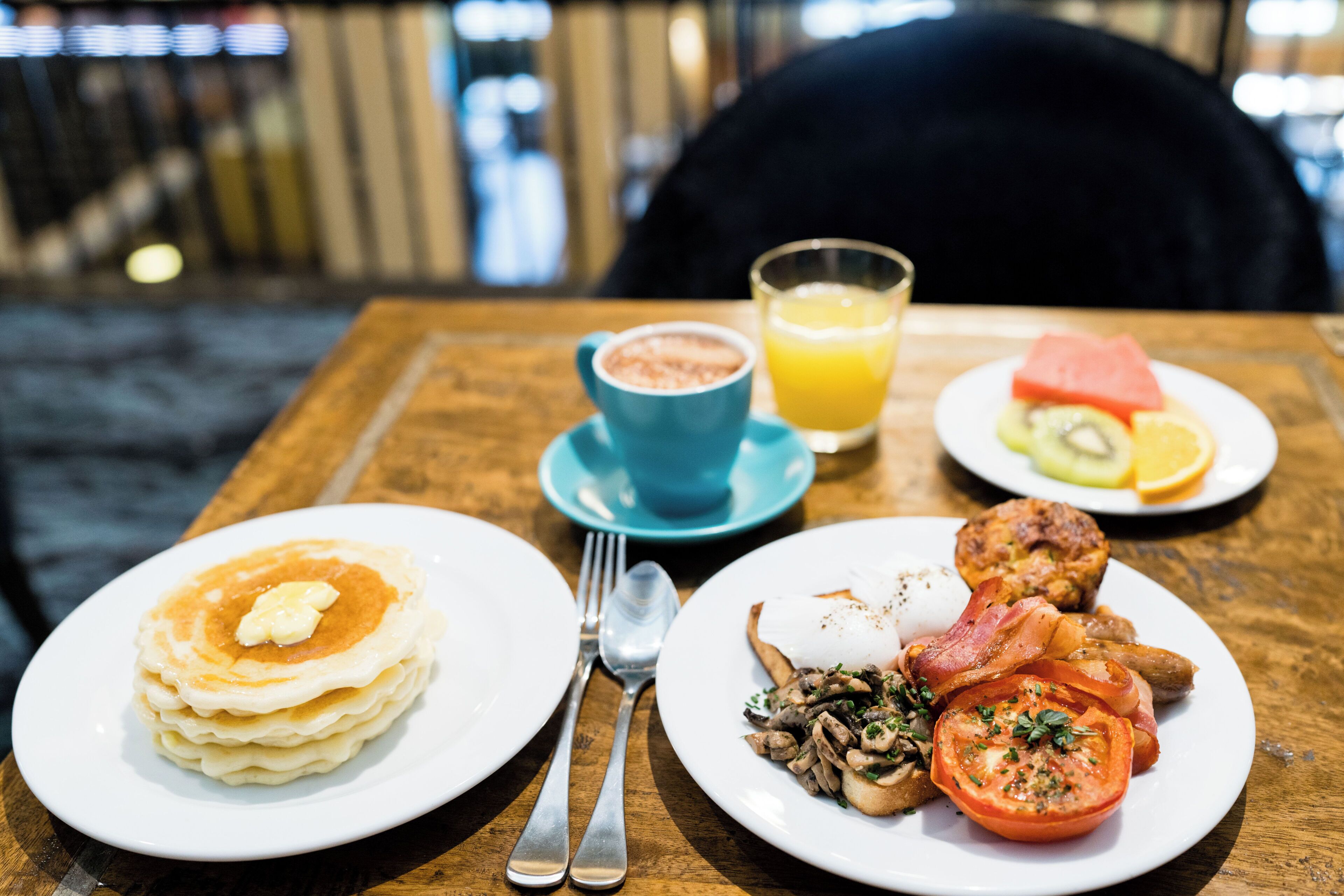 En frukostuppsättning med pannkakor, ägg, bacon och ett glas apelsinjuice på ett träbord.