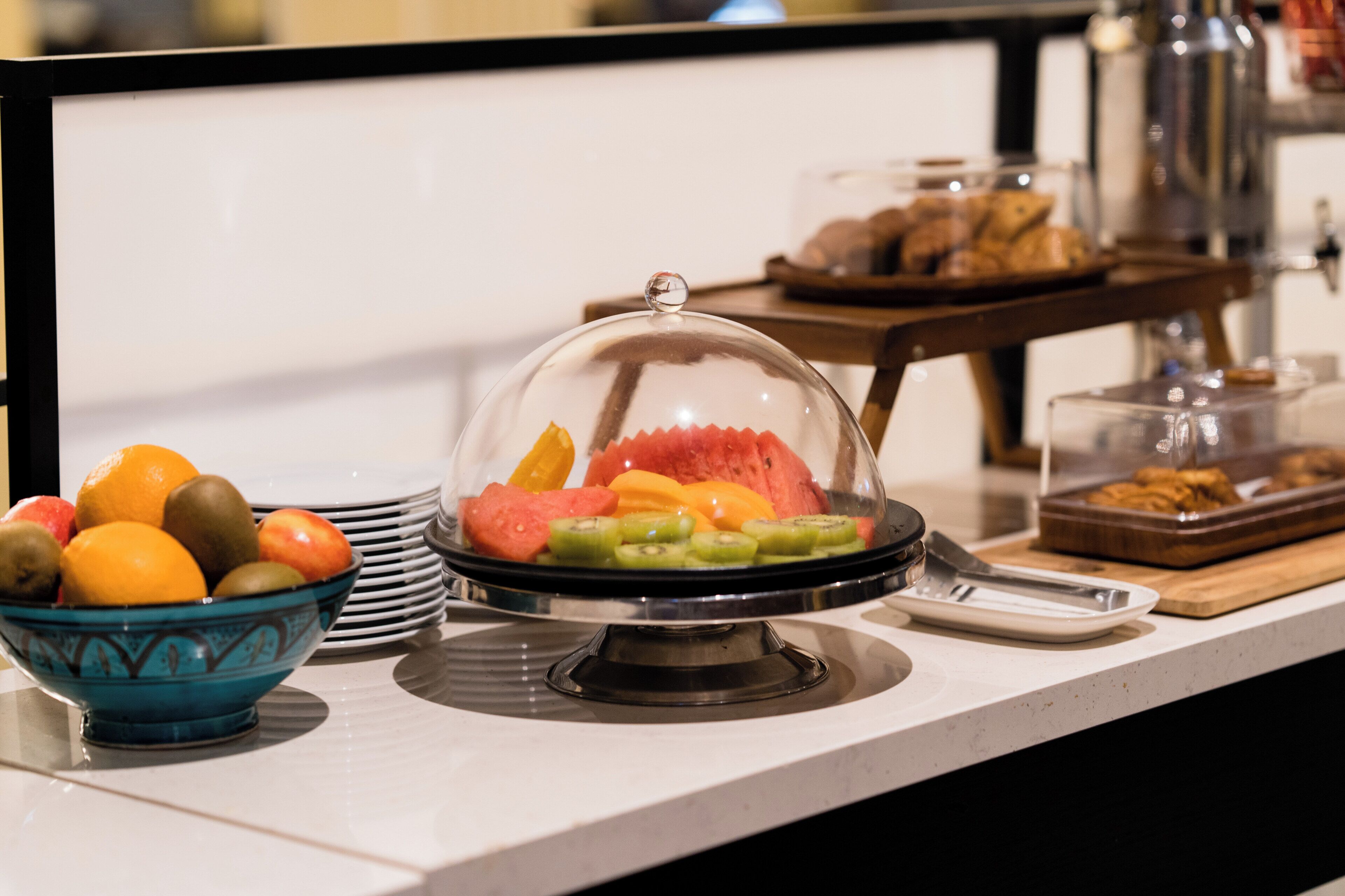 A breakfast buffet with a fruit bowl, a covered fruit display, and a tray of pastries.