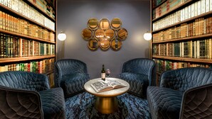 Una biblioteca con estanterías, una mesa redonda con una botella y un vaso, y dos sillones tapizados.