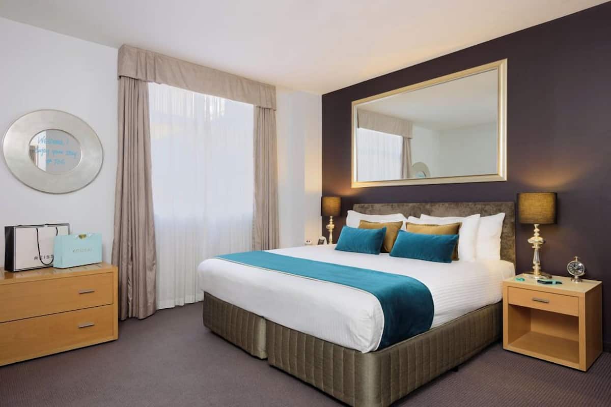 Quarto de hotel com uma cama grande, uma mesa de cabeceira com abajur, cômoda, espelho redondo e uma janela com cortinas.