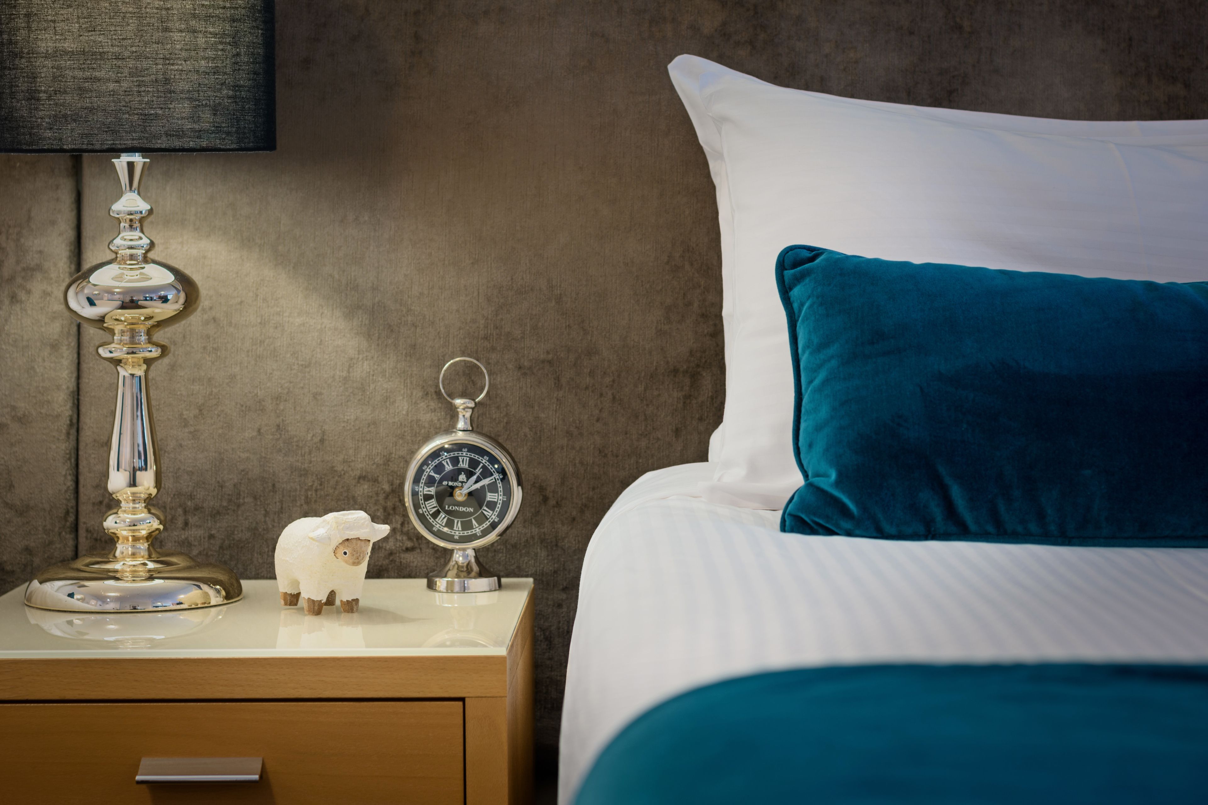A neatly made bed with a teal pillow and a white pillow, a bedside table with a lamp and a clock, and a wooden nightstand with a drawer.