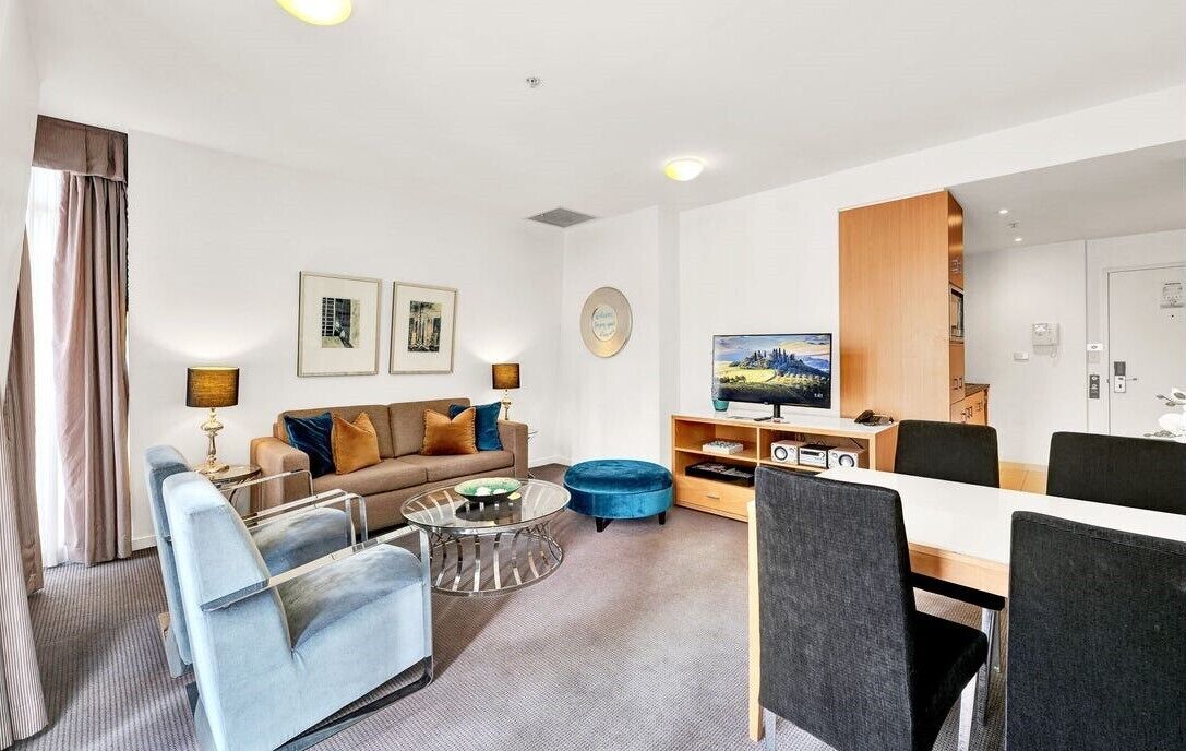 A modern hotel room with a sofa, armchairs, a dining table, and a TV.