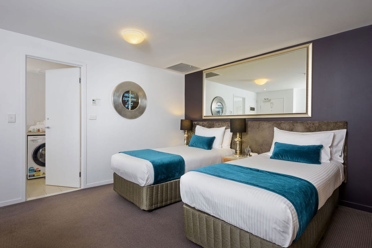 Quarto de hotel com duas camas, um espelho grande, um sinal de boas-vindas e uma área de lavanderia visível através de uma porta aberta.