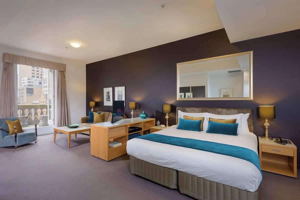 Quarto de hotel com uma cama grande, uma escrivaninha, uma cadeira e uma janela com cortinas.