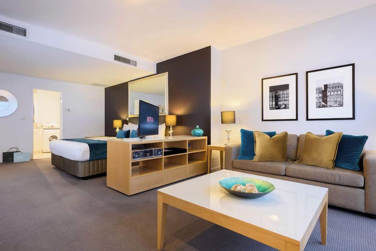 Quarto de hotel moderno com cama, sofá, mesa de centro, TV e um espelho grande.
