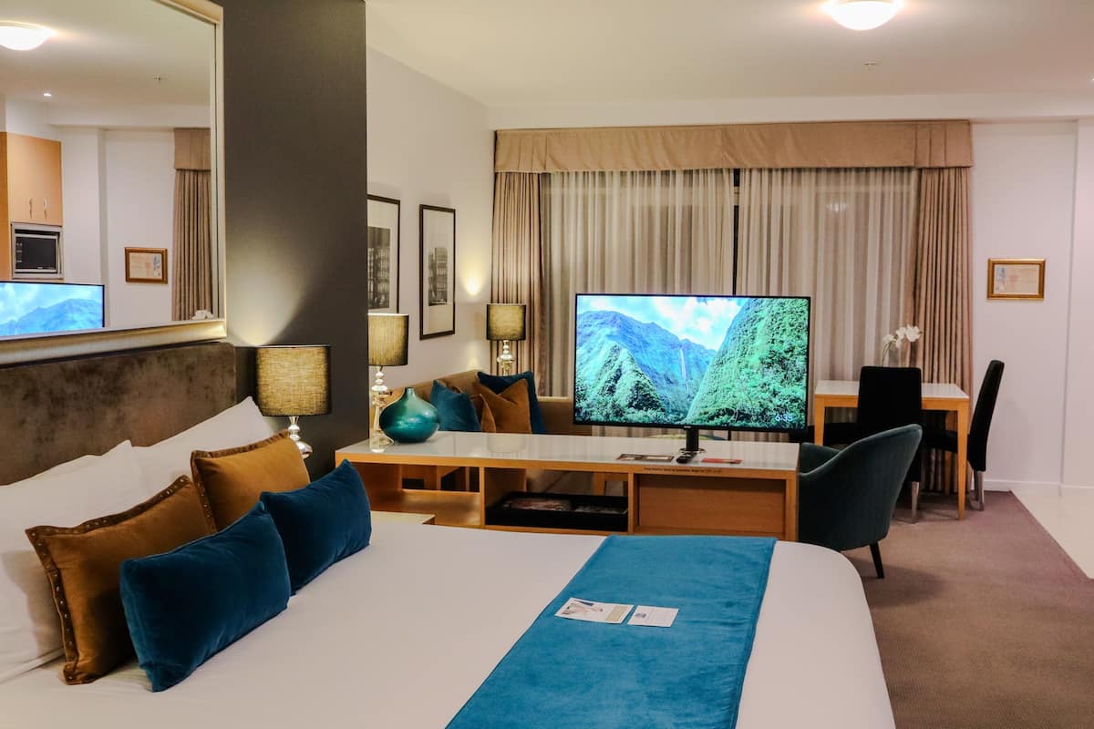 Quarto de hotel com cama, escrivaninha, televisão e área de refeições.