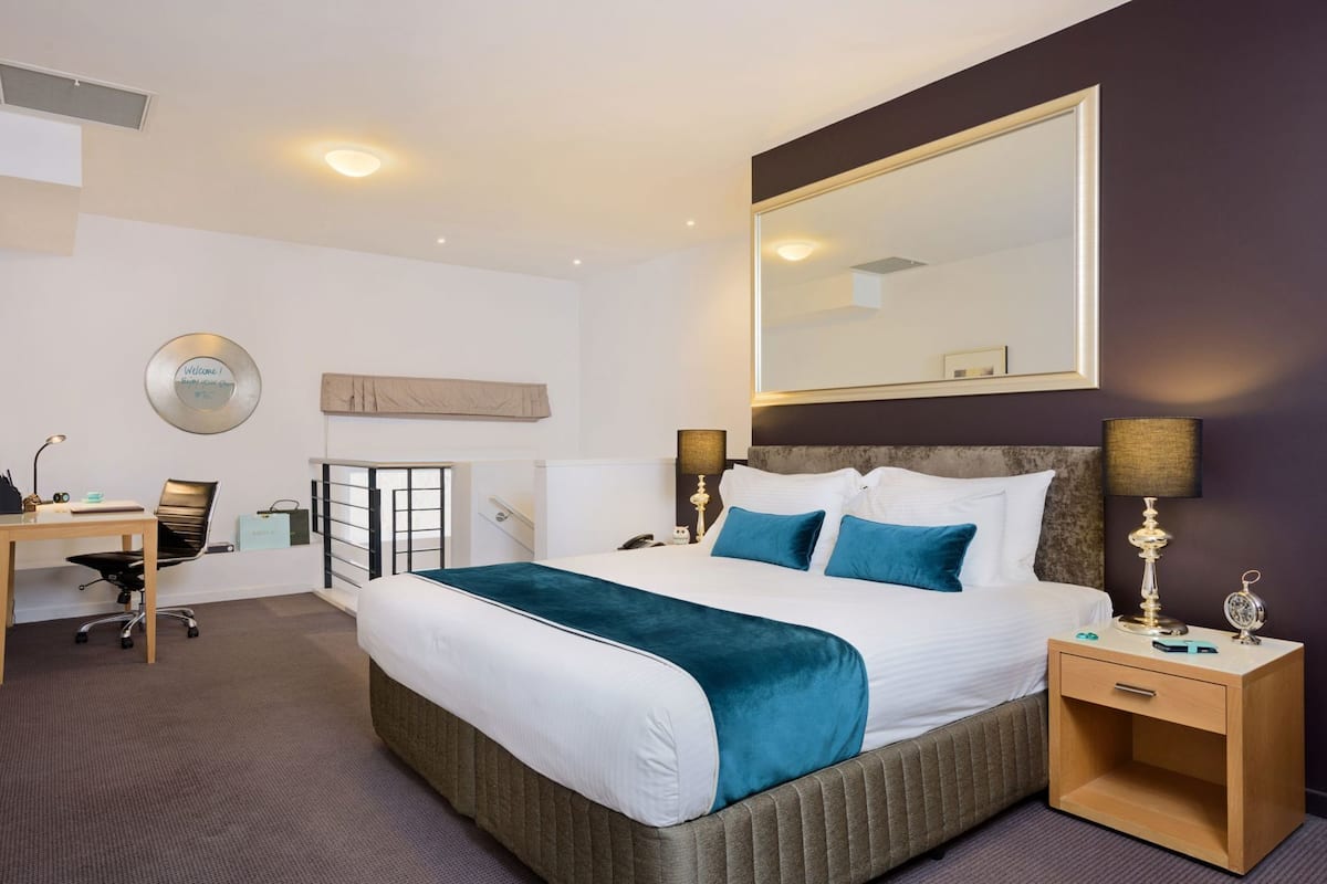 Quarto de hotel com cama, mesa de cabeceira, escrivaninha, cadeira e um espelho grande.