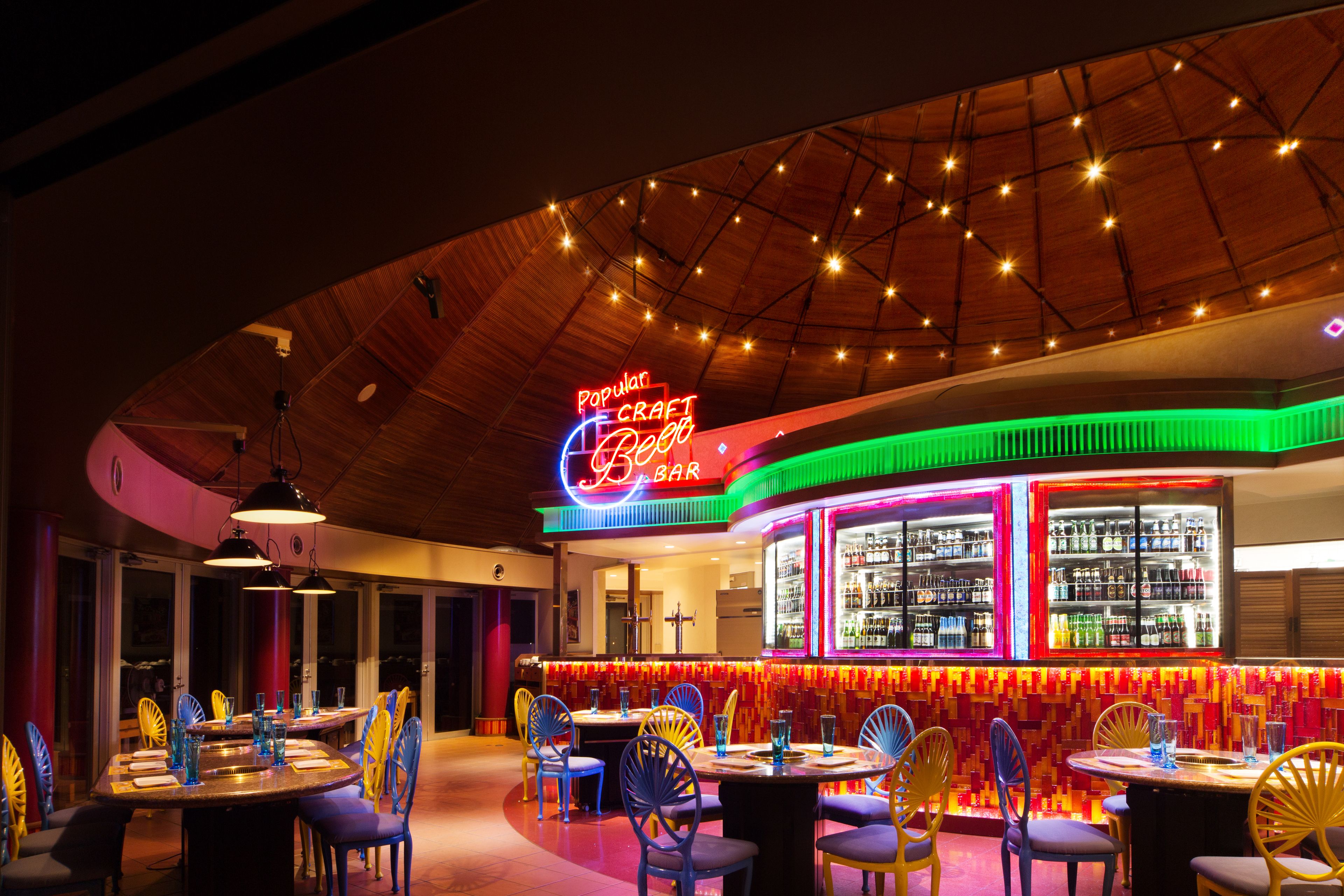 A bar with neon signage, wooden ceiling, and colorful chairs.