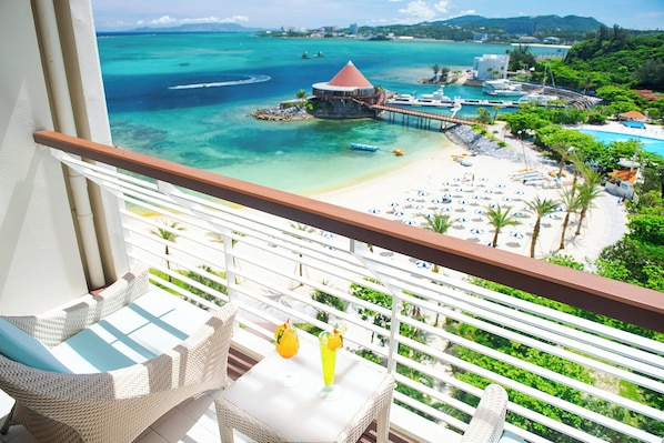 Un balcón con mobiliario de mimbre y vistas a una playa con muelle, sombrillas y piscina.