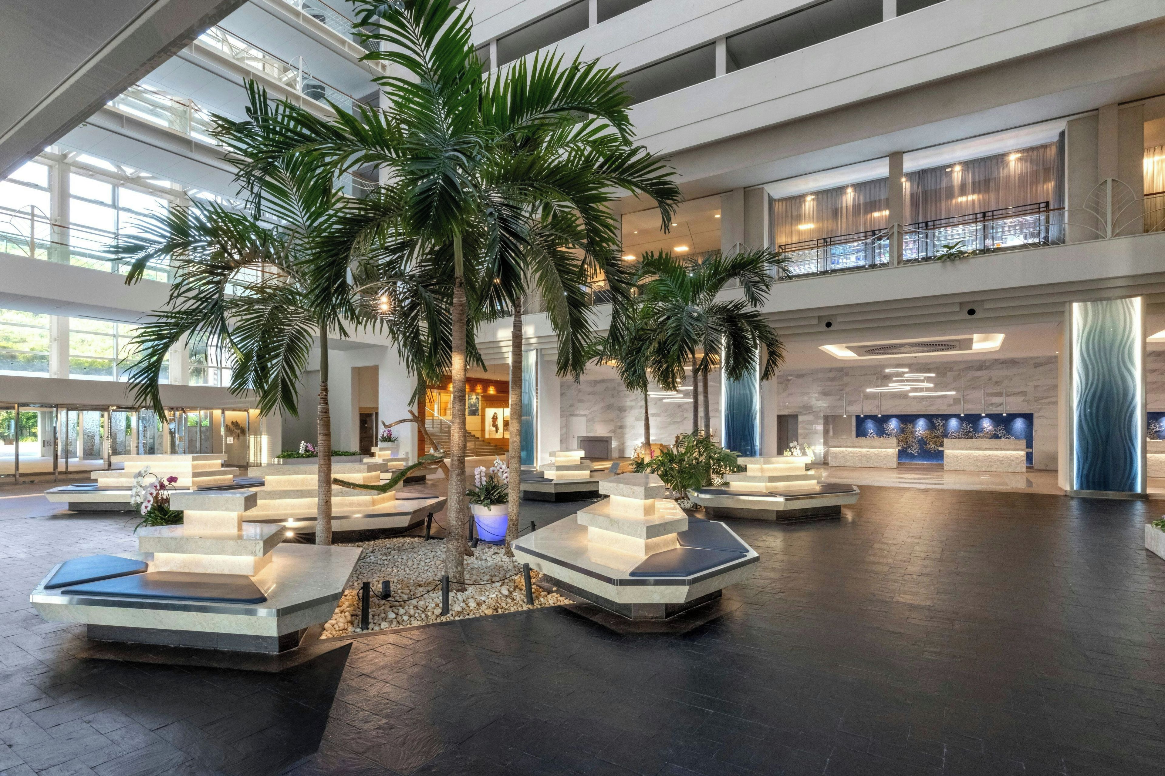 A spacious lobby with multiple seating areas, palm trees, and a modern reception desk.