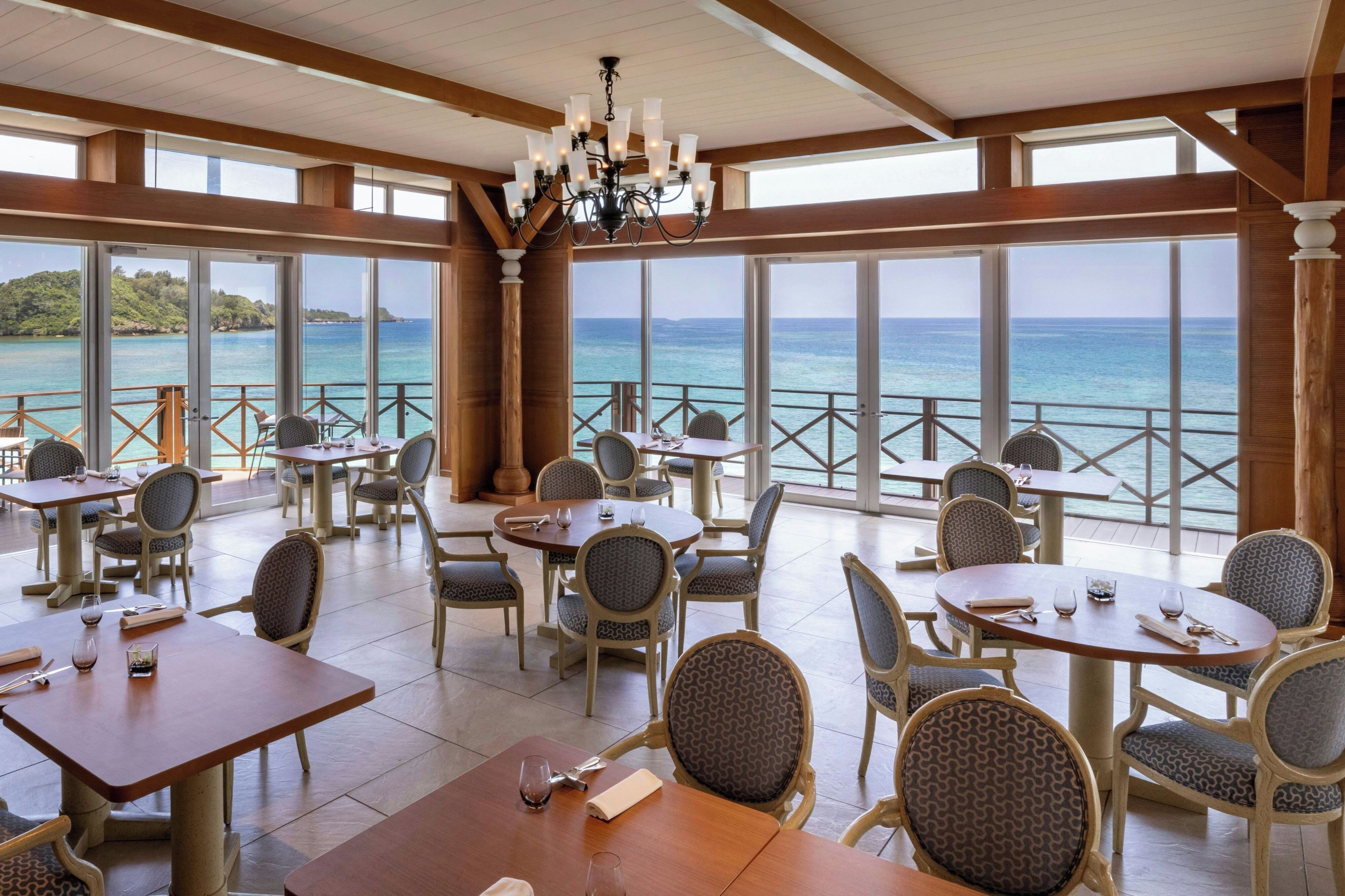 Un restaurante con mesas y sillas dispuestas para comer, amplias ventanas que ofrecen vista al mar y un candelabro colgando del techo.