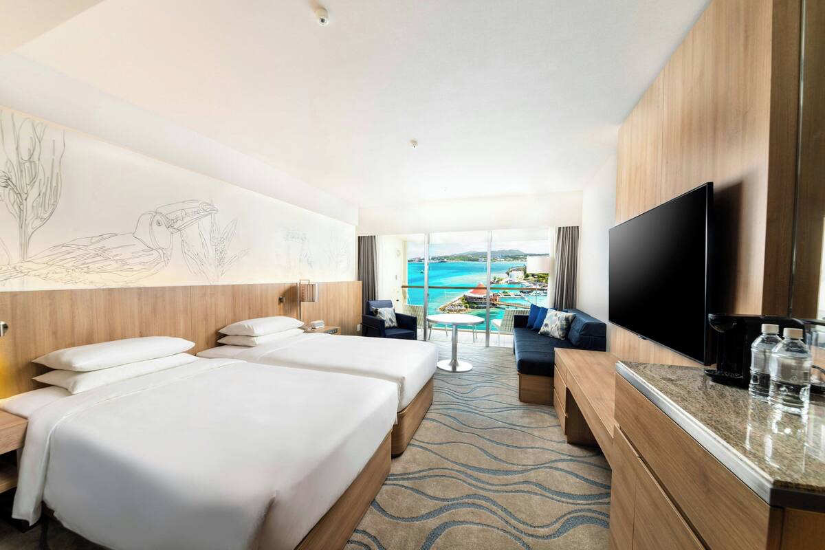 Habitación de hotel con dos camas, un televisor de pantalla plana, una zona de comedor con vistas al mar y un ventanal grande.