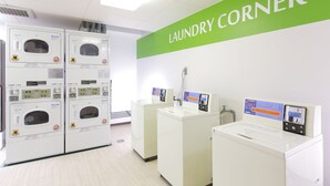Une buanderie équipée de plusieurs machines à laver et de plusieurs sèche-linge, une enseigne indiquant «Coin laverie», et un compteur d’eau.