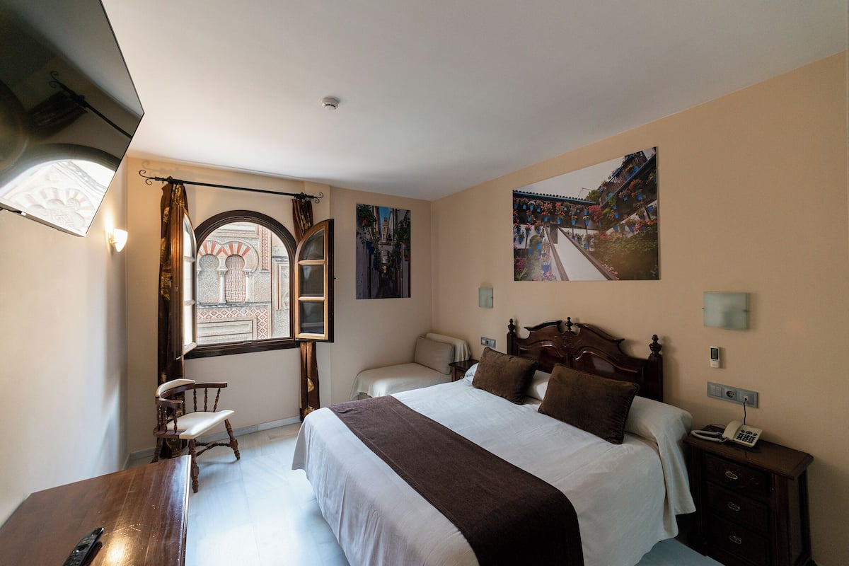 Ein Hotelzimmer mit einem großen Bett, einem Nachttisch aus Holz, einem Schreibtisch und einem Stuhl. Ein Fenster bietet Blick auf ein Gebäude, und an der Wand hängt ein Gemälde.