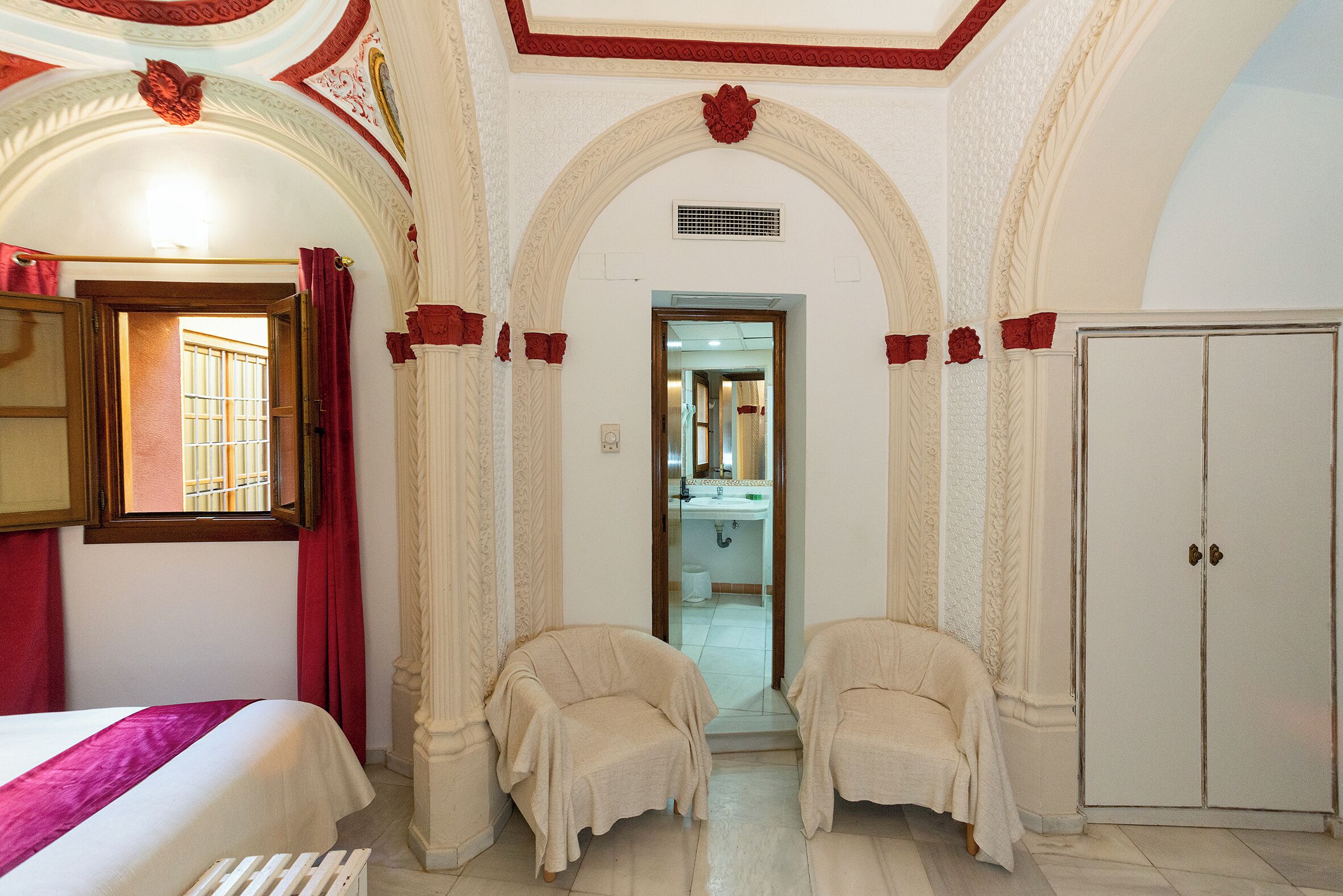 Una habitación de hotel con una cama, dos sillones, un armario y un baño visible a través de un arco.
