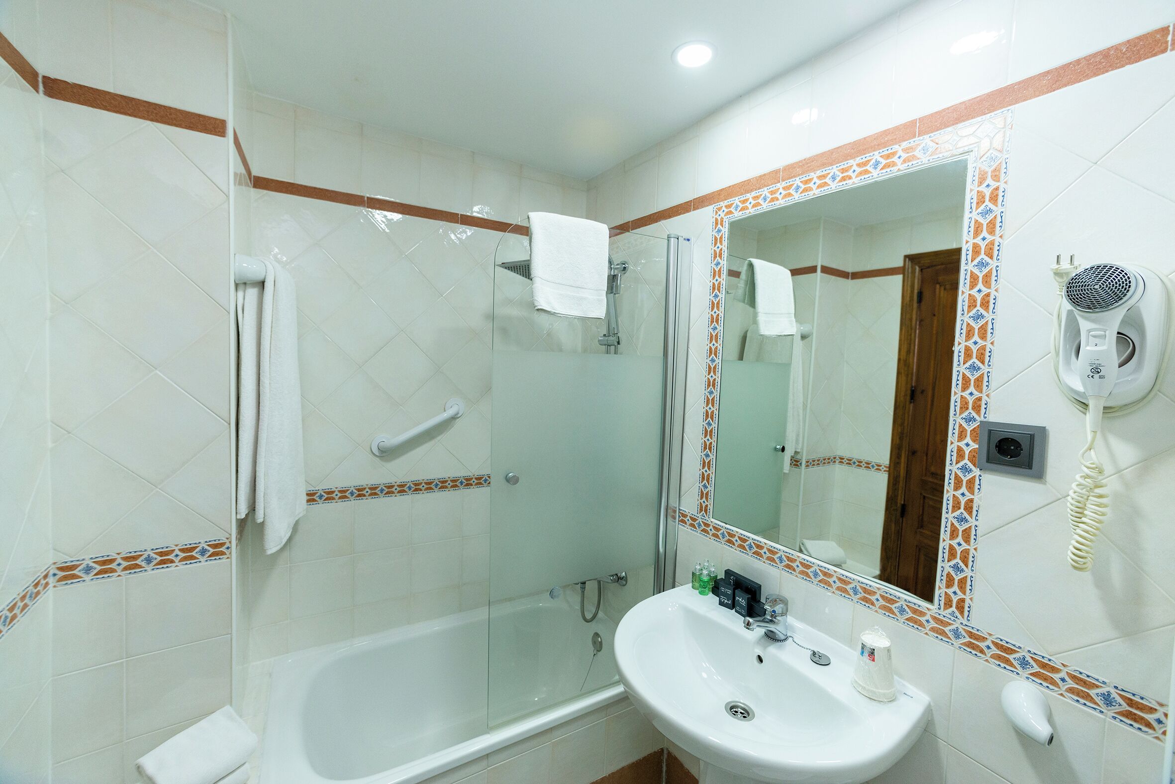 A bathroom with a glass-enclosed shower, a white sink with a mirror above it, and a hairdryer on the wall.