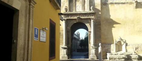 Una calle estrecha con un edificio amarillo a la izquierda y un arcoado de piedra decorativo que conduce a un edificio más grande.