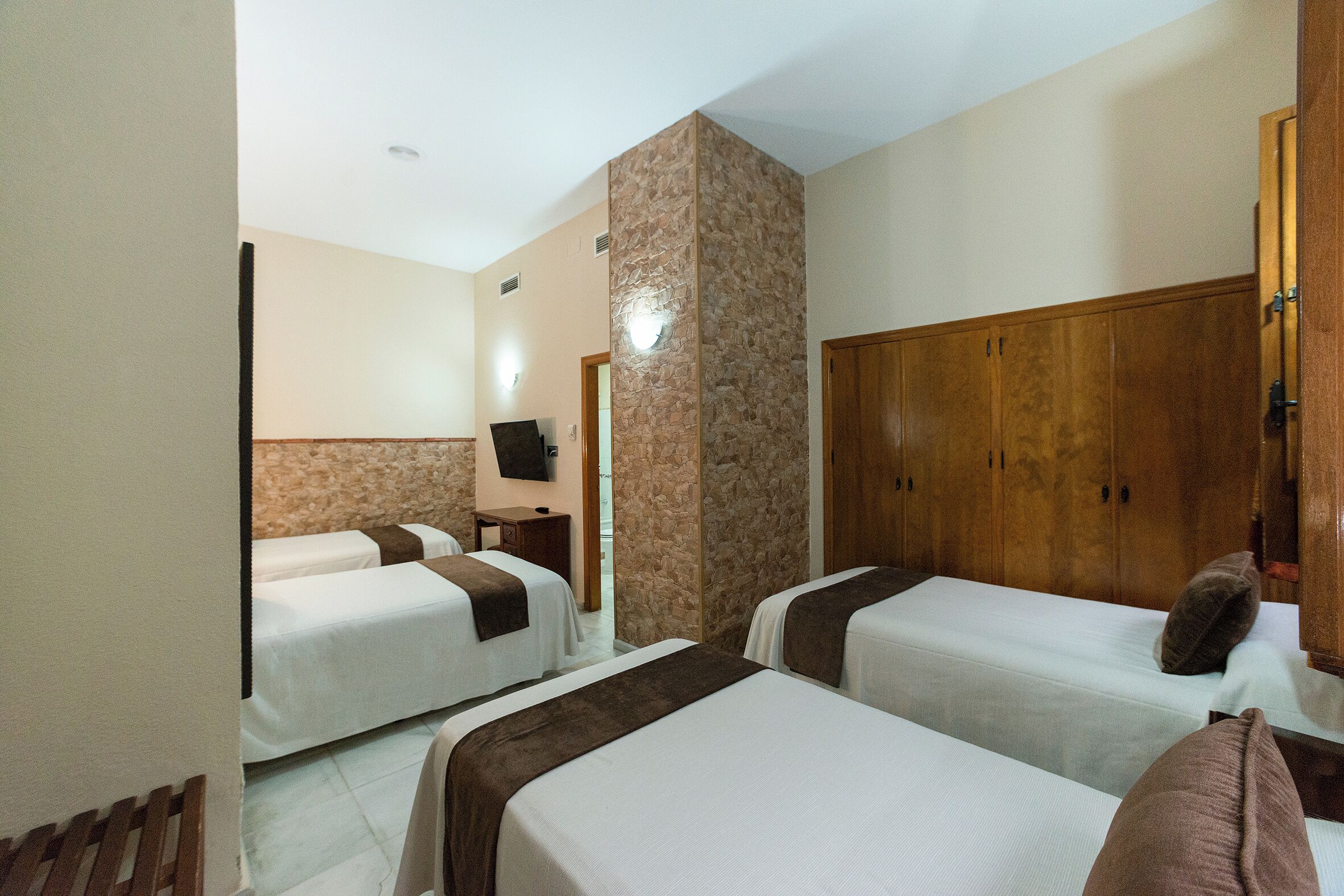 A hotel room with two single beds, a wooden wardrobe, a television on the wall, and a stone accent wall.