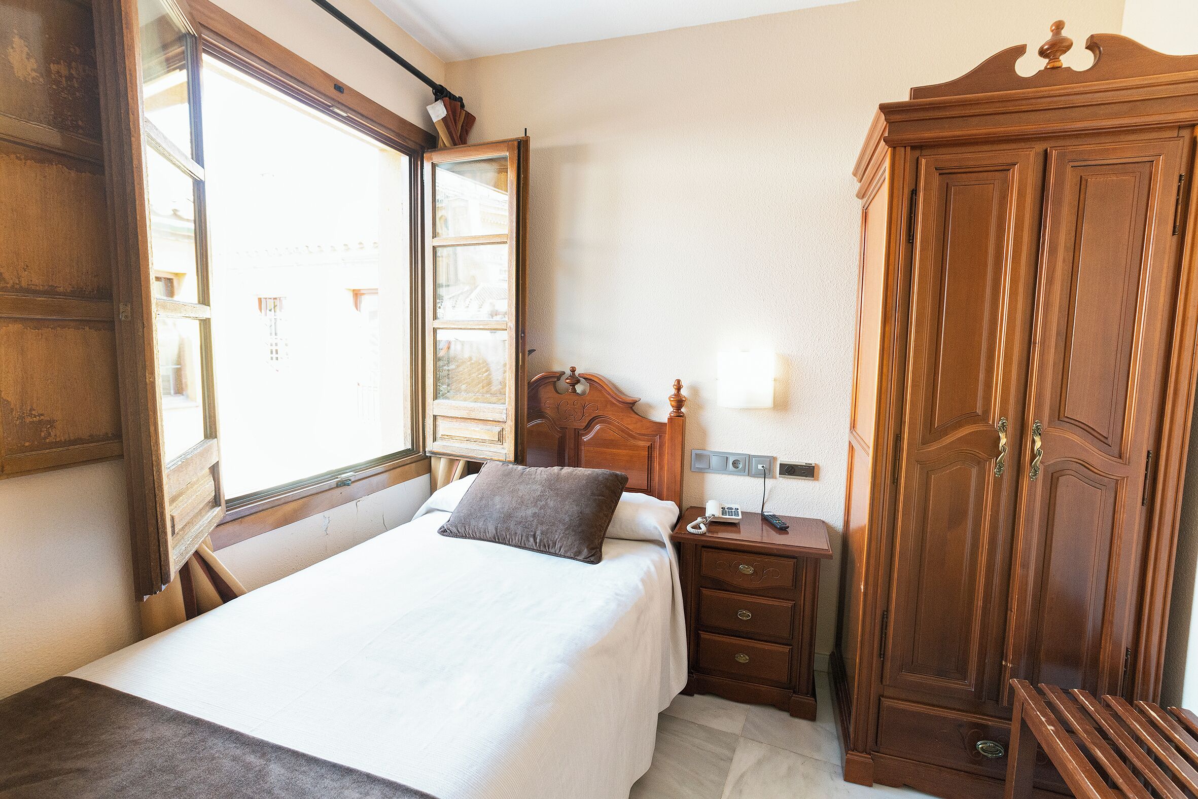 A hotel room with a bed, a wooden wardrobe, a bedside table with a lamp, and a window with curtains.