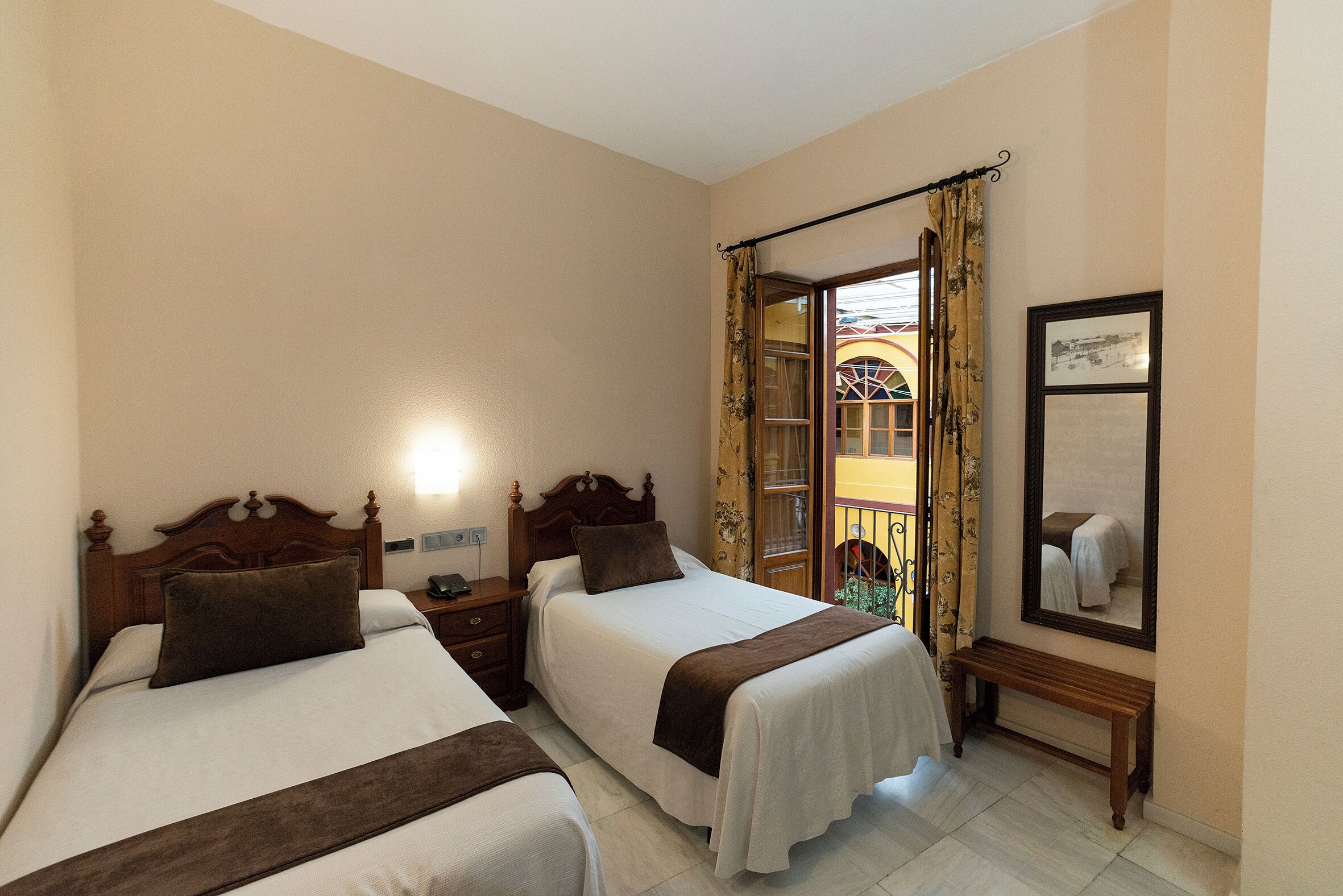 Quarto de hotel com duas camas, cabeceira de madeira, mesa de cabeceira, um banquinho, um espelho e uma janela com cortinas.