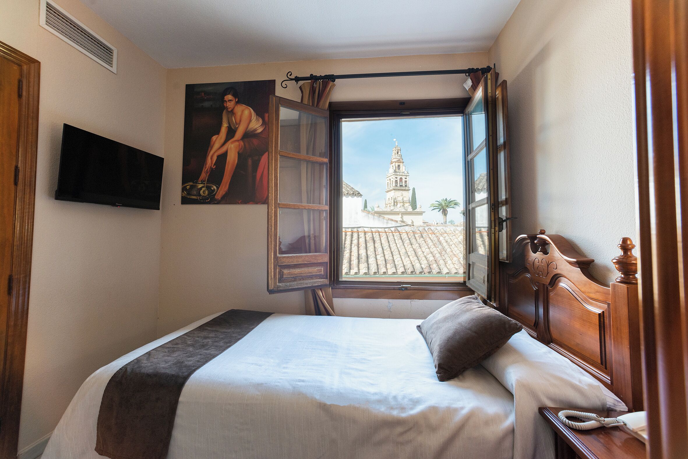 A bedroom with a bed, a painting, a television, and a window with a view of a building.