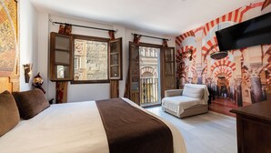 Une chambre d’hôtel avec un grand lit, un canapé, un téléviseur fixé au mur et une vue sur une cour intérieure agrémentée d’arcades.