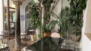 Un patio interior con mesas y sillas de metal, rodeado de plantas en macetas y un ventanal grande.