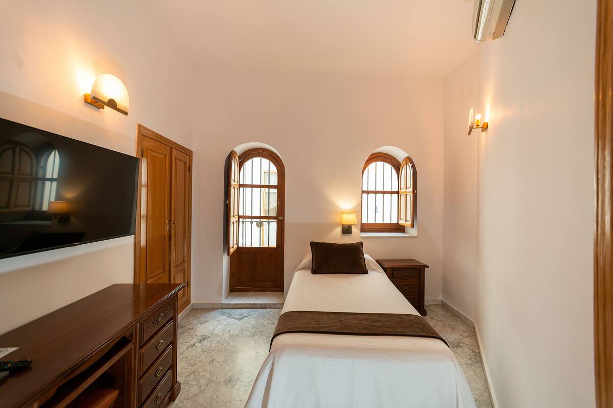 A bedroom with a bed, a TV, wooden cabinets, and two windows with arched tops.