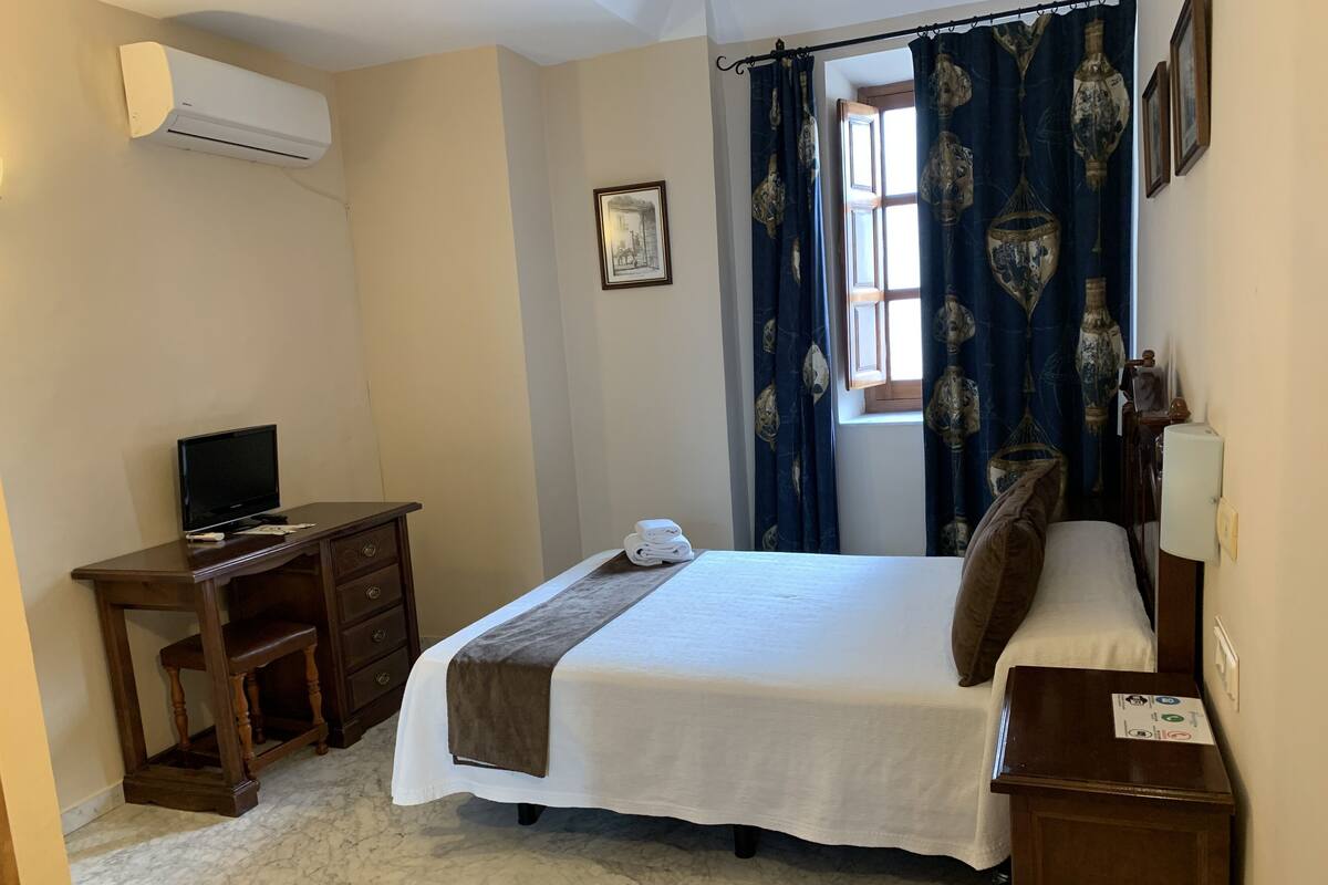 A bedroom with a bed, a desk with a computer, a TV, and a window with curtains.